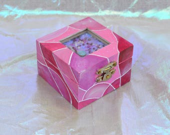 May include: A small, square, wooden jewelry box painted in shades of pink. The box features a geometric design with a clear, glass-like top. A gold-colored clasp secures the box. The interior is visible through the top, filled with small beads.