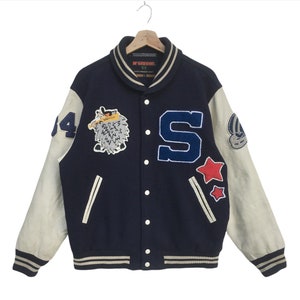 Könnte beinhalten: Eine marineblaue Varsity-Jacke mit weißen Lederärmeln. Die Jacke hat ein großes "S" auf dem Rücken, ein Comic-Tier auf der linken Brust und einen Stern auf dem rechten Ärmel.