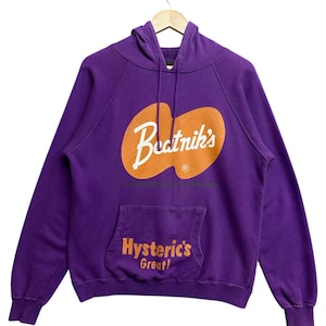 May include: A purple hoodie with a kangaroo pocket and a drawstring hood. The front features an orange design with the text "Beatnik's" and "Hysteric's Great!" in white and orange. The sweatshirt is hanging on a wooden hanger.