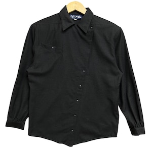 May include: Black long-sleeve shirt with a button-down front and a collared neckline. The shirt features a single chest pocket and a small label near the collar. The shirt is hanging on a wooden hanger.