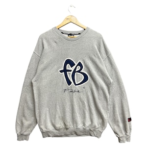 以下が含まれることがあります： 前面に大きな青と白の「fB」ロゴ、下にブランド名「Fubu」が入ったグレーのスウェットシャツ。スウェットシャツは木製のハンガーに吊るされています。