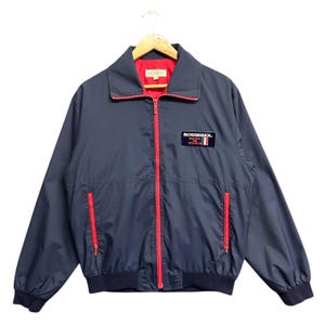 May include: Navy blue jacket with a red zip and trim. The jacket features a collar and two front pockets with red zips. A patch on the chest reads "ROSSIGNOL ÉQUIPE DE FRANCE". The jacket is suspended from a wooden hanger.