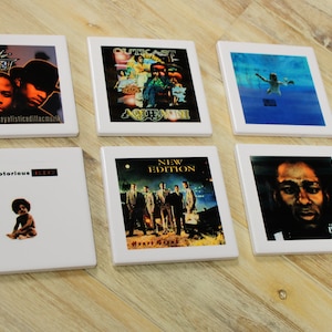 May include: Six square ceramic coasters with album art from various hip-hop artists. The coasters feature album covers from Outkast, The Notorious B.I.G., and others. The coasters are white with a glossy finish and are arranged on a wooden surface.