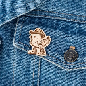Può includere: Spilla in legno raffigurante un pollo dei cartoni animati con cappello e stivali da cowboy. La spilla è attaccata a una giacca di jeans blu. La spilla è marrone chiaro con dettagli incisi. La giacca ha bottoni metallici. La spilla è sulla tasca.