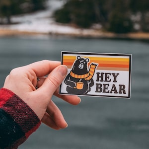 May include: A sticker with a cartoon bear wearing a scarf and holding a mug. The sticker has a yellow, orange, and brown color scheme and says "Hey Bear" in white text.
