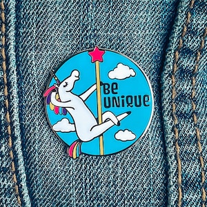 May include: A blue and white enamel pin featuring a unicorn riding a pole with the text "Be Unique".