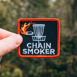 May include: A black and red embroidered patch featuring a disc golf basket and the text "Chain Smoker". The basket is on fire.