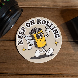 May include: A round, beige sticker with the text "Keep on Rolling" in black lettering. A cartoon film roll with a smiling face is walking to the right. The sticker is on a wooden surface.