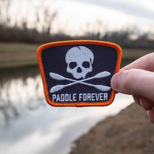 May include: A black and white embroidered patch with a skull and crossbones design. The skull has a dripping effect and the crossbones are paddles. The patch has an orange border and the text "PADDLE FOREVER" is written below the skull and crossbones.