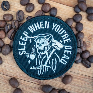 Peut inclure: Patch brodé noir avec un crâne blanc tenant une tasse de café et le texte "SLEEP WHEN YOU'RE DEAD".
