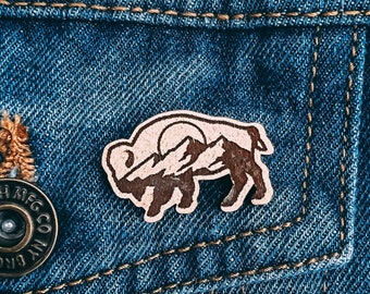 May include: A brown leather patch with a silhouette of a bison standing in front of a mountain range. The patch is pinned to a blue denim jacket.