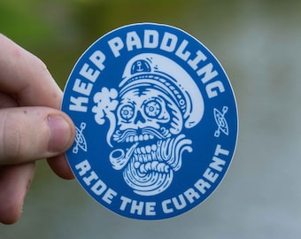 May include: A blue and white round sticker featuring a skull wearing a captain's hat and smoking a pipe. The text on the sticker reads "Keep Paddling Ride The Current".