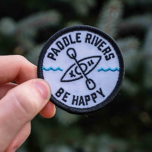 Peut inclure: Un écusson brodé blanc avec des points noirs. L'écusson représente un canoë avec des pagaies, des vagues bleues et le texte "Paddle Rivers Be Happy".