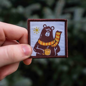 May include: A brown bear wearing a yellow and orange scarf holds a brown mug with a white rim. The bear is on a white background with a yellow sun in the top left corner. The image is surrounded by a brown border.