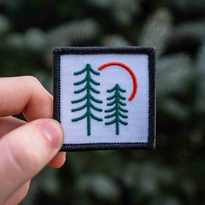 Könnte beinhalten: Ein weißer Patch mit schwarzem Rand zeigt ein gesticktes Design mit zwei grünen Bäumen und einem roten Halbkreis. Der Patch ist perfekt, um Ihren Besitztümern einen Hauch von Natur zu verleihen.