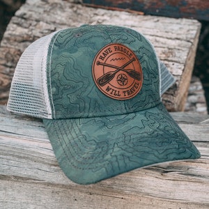 Op de afbeelding: Een groene camouflage trucker cap met een bruin leren patch waarop "Have Paddle Will Travel" staat met een kompas en gekruiste peddels.