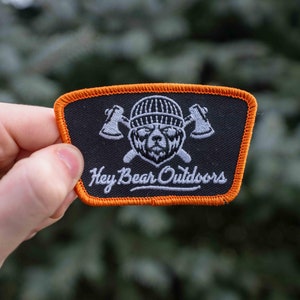 May include: Black embroidered patch with an orange border. The patch features a white bear wearing a hat with crossed axes behind it. The text "Hey Bear Outdoors" is written in white cursive below the bear.
