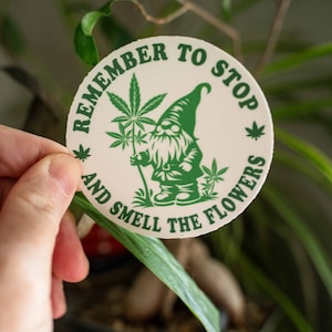 May include: A white round sticker with green text and a green cartoon gnome holding a marijuana plant. The text reads "Remember to stop and smell the flowers".