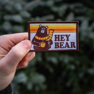 May include: A brown bear wearing a yellow and orange scarf holds a cup of coffee. The bear is on a white background with the text "HEY BEAR" in brown letters.
