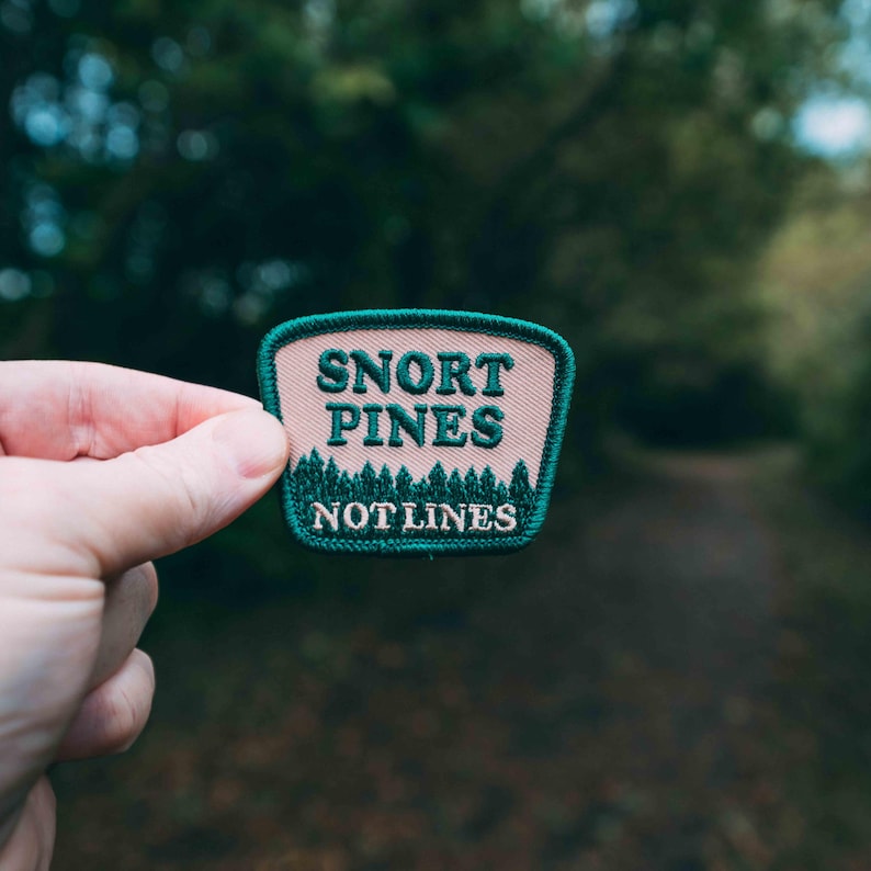 May include: A pink and green embroidered patch with the text "Snort Pines Not Lines".