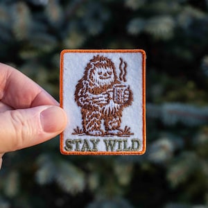 Könnte beinhalten: Gestickter Aufnäher mit einem braunen Bigfoot-Design, das einen Becher mit Dampf hält. Der Aufnäher hat einen orangefarbenen Rand und die Worte "STAY WILD" in Grün und Orange. Der Hintergrund ist eine verschwommene Ansicht von grünem Laub.