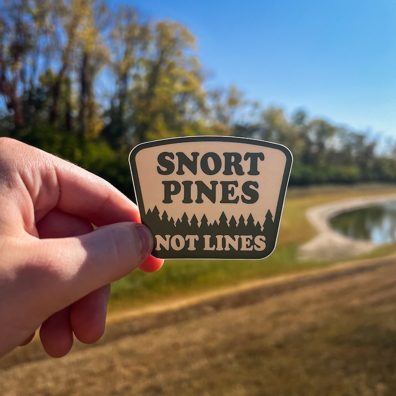 May include: A rectangular sticker with a tan background and dark green text that reads "SNORT PINES NOT LINES." The sticker has a forest silhouette at the bottom. The sticker is held in front of a blurred outdoor scene.