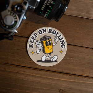 Puede incluir: Una pegatina redonda con fondo beige y el texto "Keep on Rolling" en negro. Una ilustraci&oacute;n de dibujos animados de un rollo de pel&iacute;cula con ojos y una sonrisa camina hacia la derecha. El rollo de pel&iacute;cula es amarillo con detalles negros. Hay dos peque&ntilde;as estrellas doradas en la pegatina.