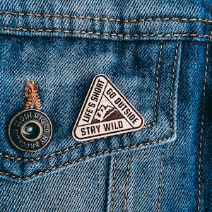 Puede incluir: Un pin de madera marrón con el texto "Life's Short Go Outside Stay Wild" en una chaqueta vaquera azul.