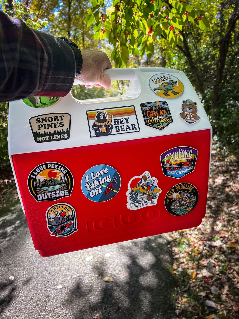 K&ouml;nnte beinhalten: Eine rot-wei&szlig;e Igloo-K&uuml;hlbox mit Aufklebern zum Thema Outdoor. Die Aufkleber zeigen Slogans wie &bdquo;Snort Pines Not Lines&ldquo; und &bdquo;I Love Yaking Off&ldquo;. Die K&uuml;hlbox wird im Freien getragen.