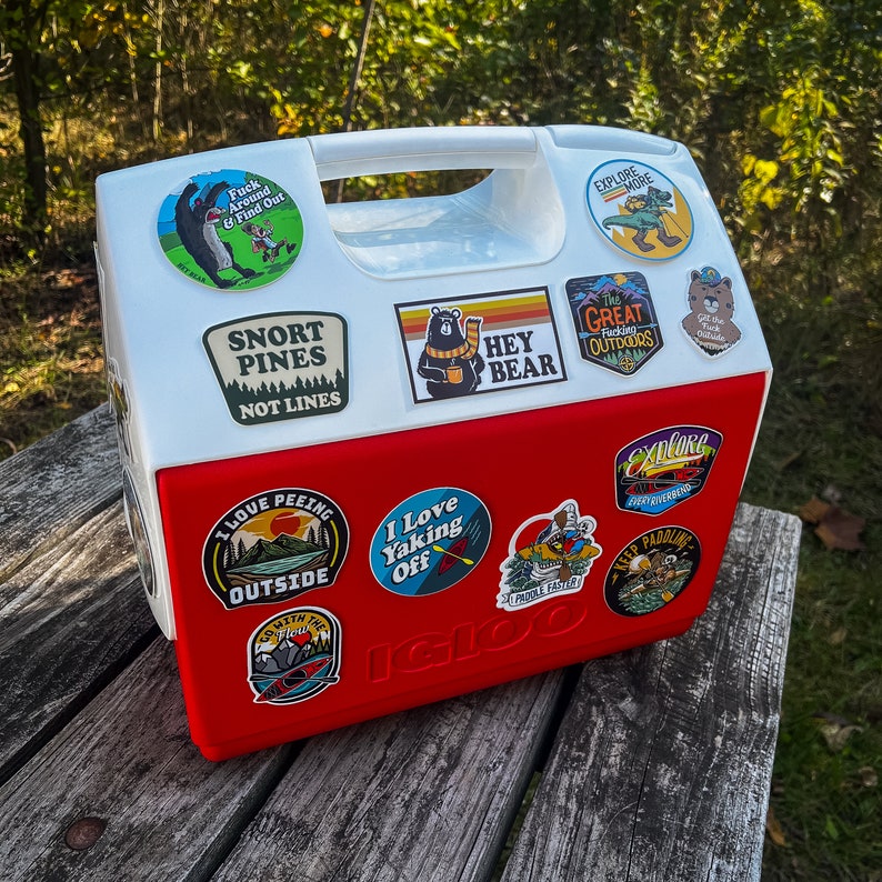 May include: A red and white Igloo cooler adorned with various outdoor adventure stickers. The stickers display slogans such as "Explore More," "Snort Pines Not Lines," and "I Love Peeing Outside." The cooler is placed on a wooden surface.