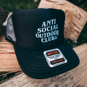 May include: Black trucker hat with the text "ANTI SOCIAL OUTDOOR CLUB" in white. The hat has a mesh back and a curved brim. A sticker on the brim reads "OTTO WORLD FAMOUS FOAM FRONT OSFM TRUCKER DURABLE SNAP CLOSURE".