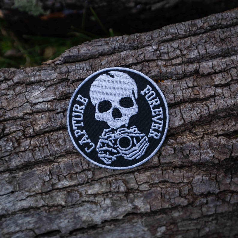 Pu&ograve; includere: Toppa ricamata in bianco e nero con uno scheletro che tiene una macchina fotografica. Il testo "CAPTURE FOREVER" &egrave; scritto intorno allo scheletro.