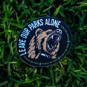 May include: A circular sticker with the text "LEAVE OUR PARKS ALONE" in white on a black background. The sticker features a brown bear with an open mouth. The words "Protect Our Parks" and a green tree are also on the sticker.