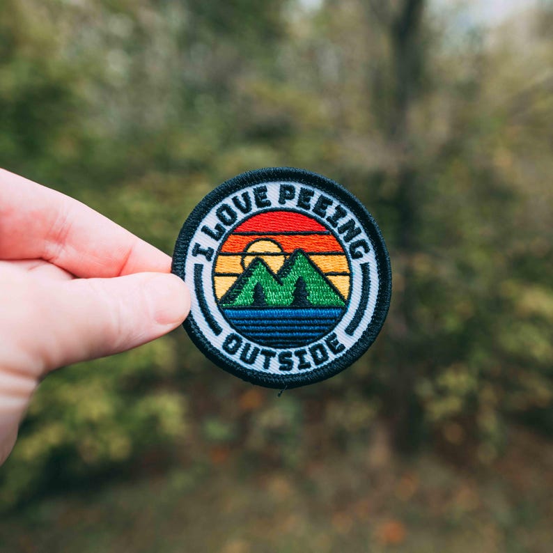 May include: A black and white embroidered patch with a rainbow sunset, mountains, and a lake. The text on the patch reads "I Love Peeing Outside".