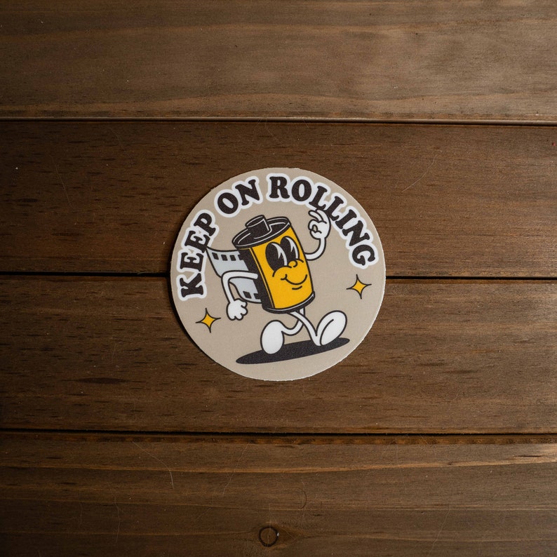 Puede incluir: Una pegatina redonda con un personaje de rollo de pel&iacute;cula de dibujos animados con el texto "Keep on Rolling" en letras marrones sobre un fondo beige.