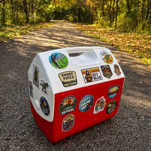 May include: A red and white Igloo cooler adorned with outdoor adventure stickers. The stickers feature phrases such as "Snort Pines Not Lines" and "I Love Yaking Off." The cooler is positioned on a paved path, with trees visible in the background.