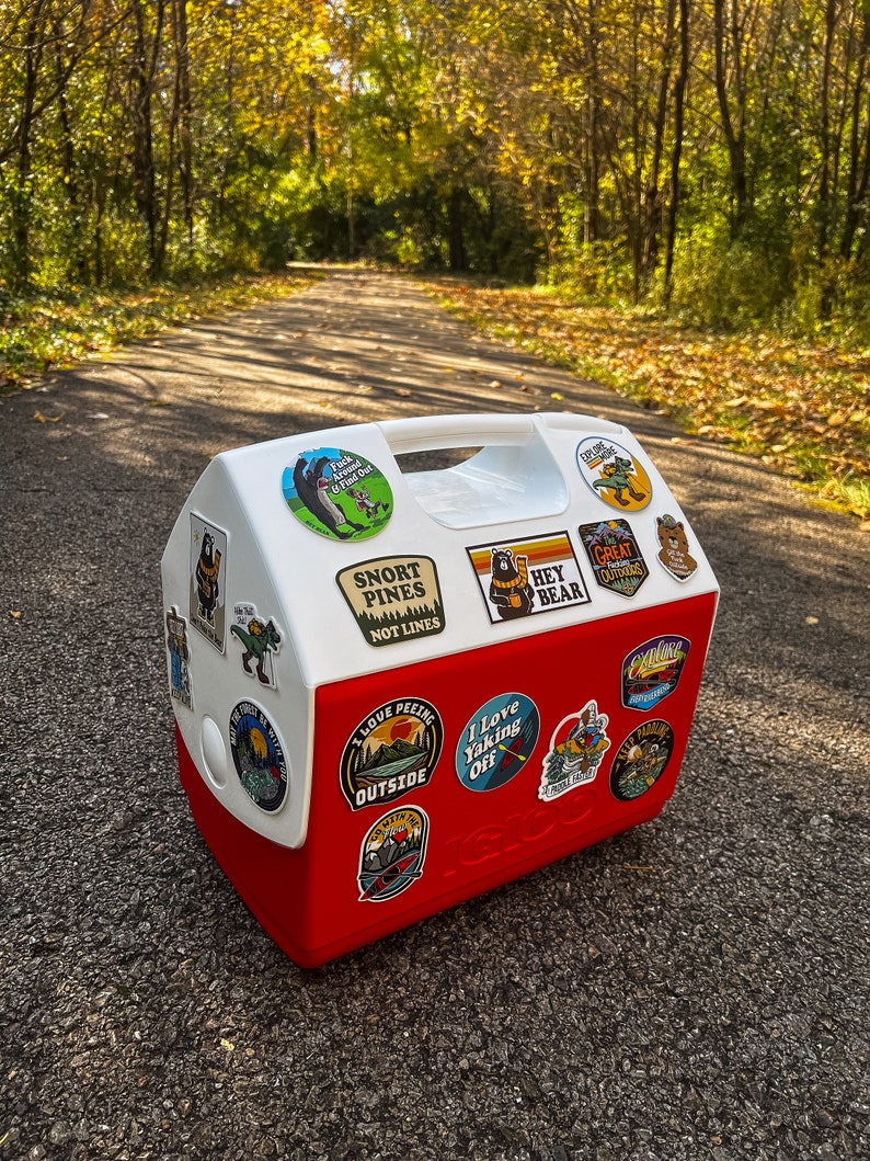 May include: A red and white Igloo cooler adorned with outdoor adventure stickers. The stickers feature phrases such as "Snort Pines Not Lines" and "I Love Yaking Off." The cooler is positioned on a paved path, with trees visible in the background.