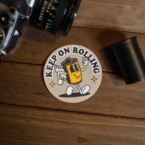 Puede incluir: Una pegatina redonda con un personaje de rollo de pel&iacute;cula de dibujos animados con el texto "Keep on Rolling" en una fuente de estilo retro.
