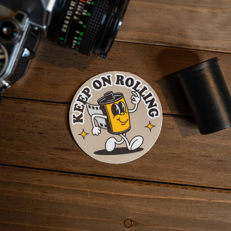 Puede incluir: Una pegatina redonda con un personaje de rollo de pel&iacute;cula de dibujos animados con el texto "Keep on Rolling" en una fuente de estilo retro.
