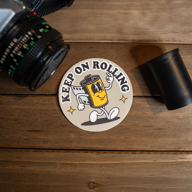 Puede incluir: Una pegatina redonda de color beige con el texto "Keep on Rolling" en letras negras. Un rollo de pel&iacute;cula de dibujos animados con una cara sonriente camina hacia la derecha. La pegatina est&aacute; sobre una superficie de madera.