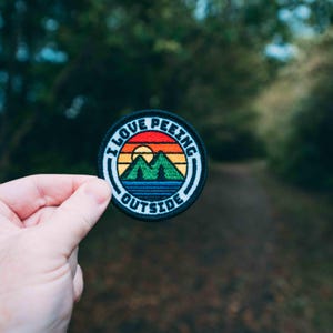 May include: A black and white embroidered patch with a rainbow sunset, mountains, and a lake. The text on the patch reads "I Love Peeking Outside".