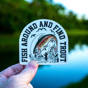 Puede incluir: Pegatina blanca con el texto "FISH AROUND AND FIND TROUT" en un diseño curvo. La pegatina presenta una colorida ilustración de una trucha saltando del agua, con un pescador y árboles en el fondo.