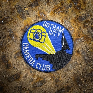 May include: A blue and yellow embroidered patch with the text "Gotham City Camera Club" and a stylised image of a camera with a flash.