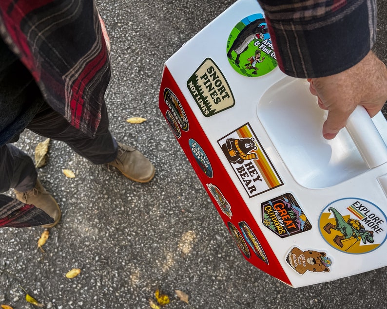 May include: A white and red cooler adorned with outdoor-themed stickers, featuring bears, mountains, and the text "HEY BEAR" and "EXPLORE MORE". The cooler is held by a person wearing a plaid shirt and grey trousers.