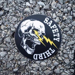 Puede incluir: Adhesivo redondo negro con una calavera blanca y un rayo amarillo que atraviesa la cuenca del ojo. Las palabras "SAFETY THIRD" están impresas en blanco alrededor de la calavera.