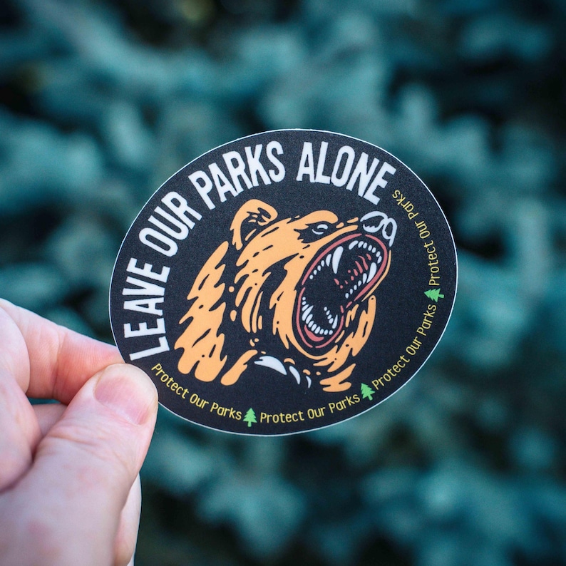 May include: A circular sticker featuring the words "LEAVE OUR PARKS ALONE" and a roaring bear illustration. The bear is orange and white, set against a black background. The words "Protect Our Parks" are also printed on the sticker.