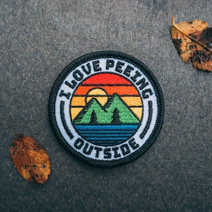 May include: A black and white embroidered patch with a sunset and mountains. The text "I Love Peeling Outside" is written around the edge of the patch.