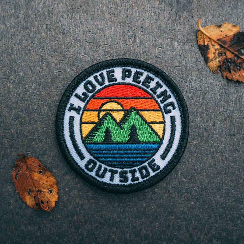 May include: A black and white embroidered patch with a sunset and mountains. The text "I Love Peeling Outside" is written around the edge of the patch.