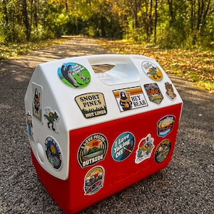 May include: A red and white Igloo cooler covered in outdoor-themed stickers. The stickers feature images of bears, mountains, and text like "Snort Pines Not Lines" and "Hey Bear." The cooler has a white handle and is sitting on a paved surface.