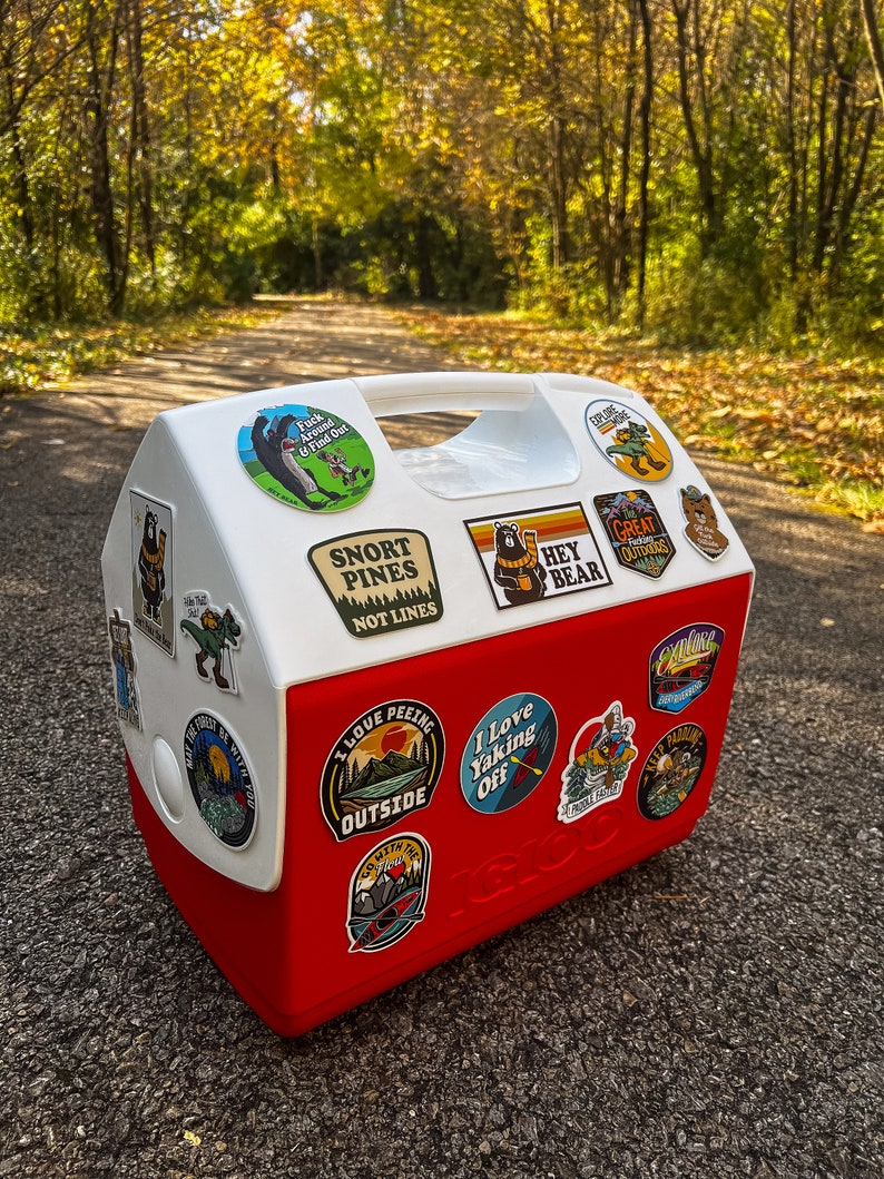 May include: A red and white Igloo cooler covered in outdoor-themed stickers. The stickers feature images of bears, mountains, and text like "Snort Pines Not Lines" and "Hey Bear." The cooler has a white handle and is sitting on a paved surface.